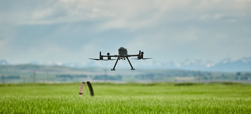 A drone hovering low over a green field, with mountains and clouds in the background.